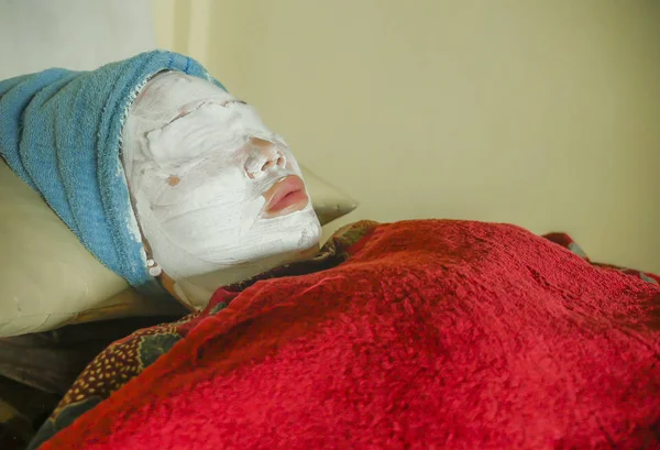 young woman with towel wrapped on head and face mask lying on bed at wellness spa receiving beauty facial treatment in well being skin care and healthy lifestyle concept