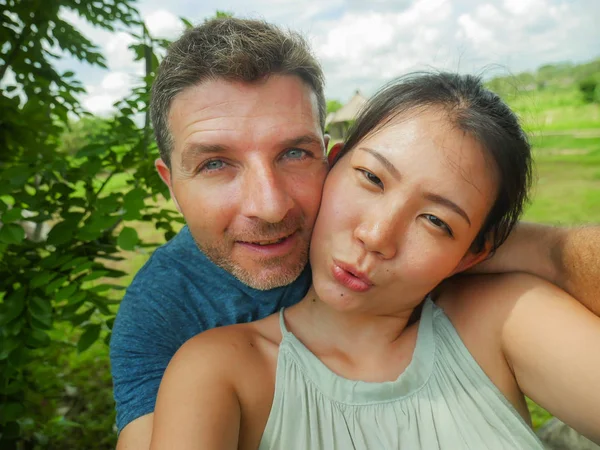 young beautiful and happy mixed ethnicity couple beautiful Asian Korean woman and white man in love taking selfie picture outdoors enjoying romantic holidays trip in tropical honeymoon vacation