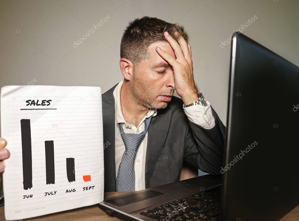 young depressed businessman working in stress at office computer desk holding notepad showing massive benefits loss and sales drop graph in corporate job problem and business crisis