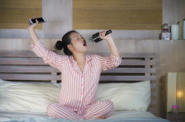 Genç mutlu ve güzel Asyalı Genç kız şarkı karaoke şarkı evde yatak holding cep telefonu yatağa heyecanlı ve neşeli pop müzik taklit oyun heyecanlı Japon yıldız