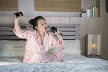 Genç mutlu ve güzel Asyalı Genç kız şarkı karaoke şarkı evde yatak holding cep telefonu yatağa heyecanlı ve neşeli pop müzik taklit oyun heyecanlı Çinli yıldız