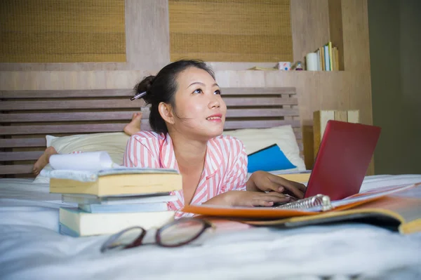 young beautiful and smart Asian American student woman at home bedroom sitting on bed studying for exam with laptop computer and textbooks smiling confident in university and education concept