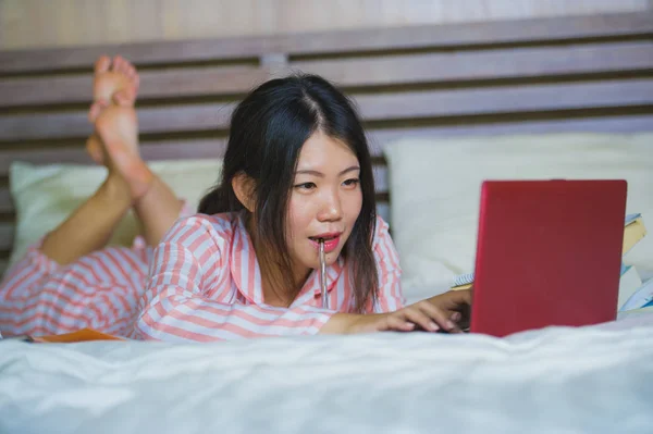 young beautiful and smart Asian Korean student girl at home bedroom sitting on bed studying for exam with laptop computer and textbooks smiling confident in university and education concept