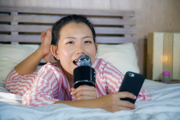 Young happy and beautiful Asian Chinese teenager girl singing karaoke song excited at home bedroom holding mobile phone playing on bed excited and cheerful emulating pop music star