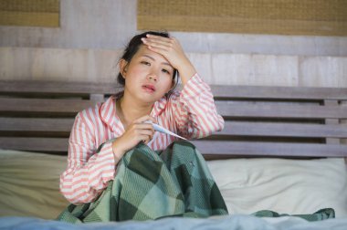 young beautiful and sweet Asian Korean girl in pajamas feeling sick at home bedroom suffering cold and flu taking temperature with thermometer in bed covered with blanket with virus infection clipart