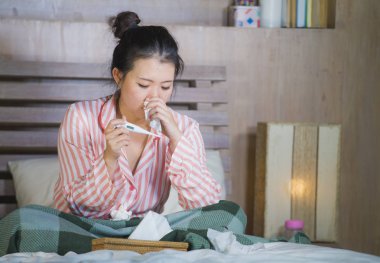 young beautiful and sweet Asian Japanese girl in pajamas sick at home bedroom suffering cold and flu taking temperature with thermometer in bed blowing her nose coughing and sneezing clipart