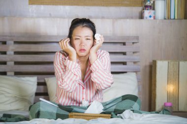 young beautiful and sweet Asian Korean woman in pajamas sick at home bedroom suffering cold and flu taking temperature with thermometer in bed blowing her nose coughing and sneezing clipart
