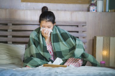young beautiful and sweet Asian Chinese woman in pajamas sick at home bedroom suffering cold and flu taking temperature with thermometer in bed blowing her nose coughing and sneezing clipart