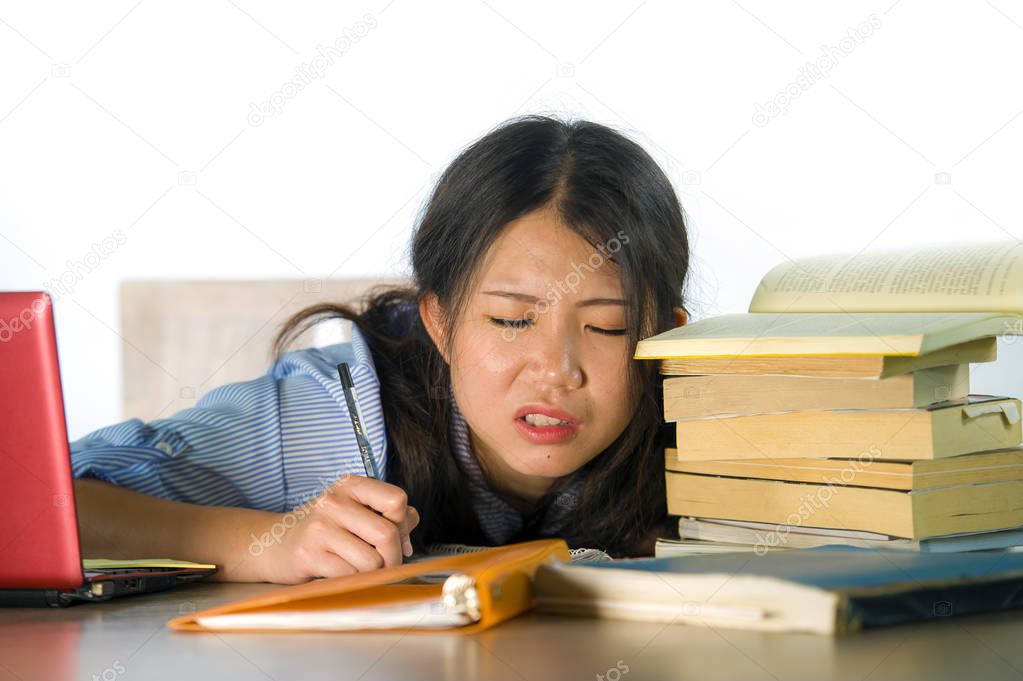 young stressed and frustrated Asian Korean teenager student working hard with laptop computer and books pile on desk overwhelmed and exhausted feeling tired and worried isolated on white