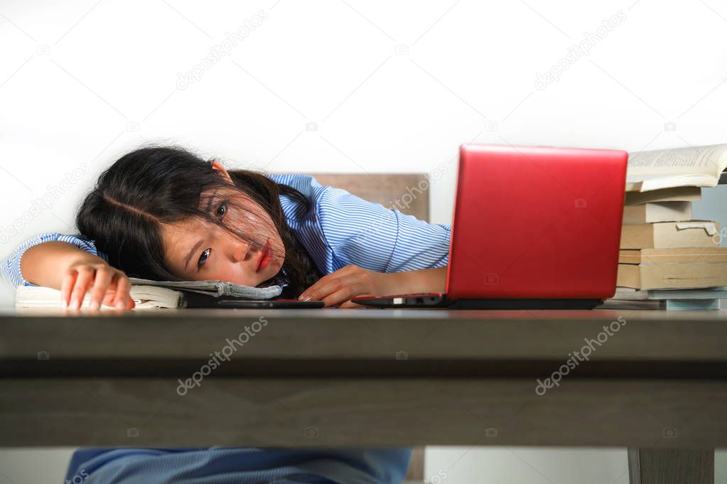 young stressed and frustrated Asian Korean teenager student working with textbooks and laptop computer on desk overwhelmed and exhausted leaning on table desperate worried about exam