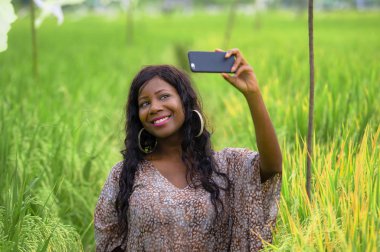 Genç mutlu ve güzel siyah Afrikalı-Amerikalı kadın şık elbise poz cool cep telefonu gülümseyen selfie pirinç alan zevk tropikal tatil de neşeli egzotik köyde fotoğrafta