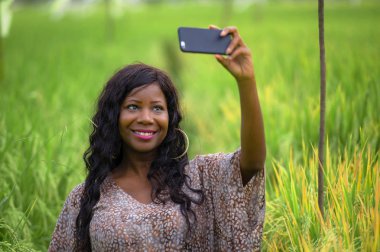 Genç mutlu ve güzel siyah afro Amerikan kadın şık elbise poz cool cep telefonu gülümseyen selfie pirinç alan zevk tropikal tatil de neşeli egzotik köyde fotoğrafta