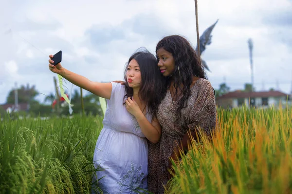 mixed ethnicity Asian Chinese girl and black African American woman taking girlfriends selfie with mobile phone together isolated at rice field enjoying exotic tropical holidays in traditional village