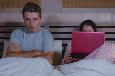 angry and frustrated husband moody in bed ignored by his workaholic wife or internet social media addict girlfriend using laptop in bed ignoring the man in couple problem and internet conflict clipart