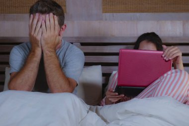 angry and frustrated husband moody in bed ignored by his workaholic wife or internet social media addict girlfriend using laptop in bed ignoring the man in couple problem and internet conflict clipart