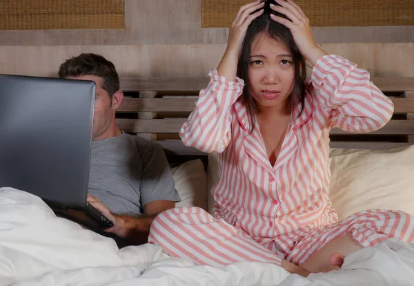 angry and frustrated Korean woman in bed ignored by workaholic husband or internet social media addict boyfriend networking with laptop in bed ignoring the woman in Asian couple problem conflict