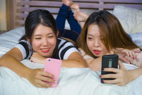 つ若い幸せとはかなりアジア韓国のガール フレンドに座って笑って 話を持っている家の寝室で楽しい女性友情でベッドの上に座って携帯電話でインターネットのソーシャル メディア アプリを使用して — ストック写真