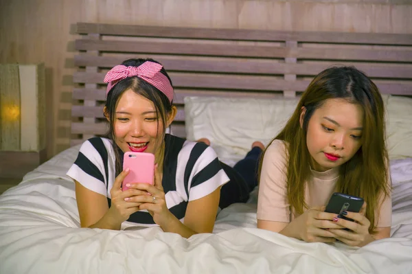 two young happy and pretty Asian Chinese girlfriends sitting at home bedroom laughing and talking having fun using internet social media app with mobile phone sitting on bed in women friendship