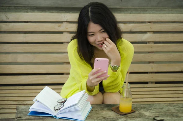 young beautiful and happy Asian Japanese woman sitting relaxed and happy at vintage wood coffee shop drinking orange juice reading novel book and using mobile phone in girl leisure weekend lifestyle