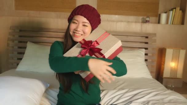 年轻快乐和美丽的亚裔美国女孩在冬天的帽子举行圣诞礼物盒与丝带微笑兴奋和欢快坐在家里的床上庆祝圣诞节收到礼物 — 图库视频影像