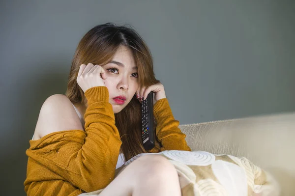 young attractive and cute scared Asian Korean woman alone at home living room couch watching horror movie or TV drama holding remote controller in fear and panic face expression