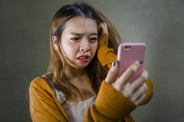 young cute and sad Asian Korean girl feeling broken heart and desperate holding mobile phone suffering relationship break up dumped via internet message isolated on dark background