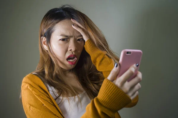 young cute and sad Asian Korean girl feeling broken heart and desperate holding mobile phone suffering relationship break up dumped via internet message isolated on dark background