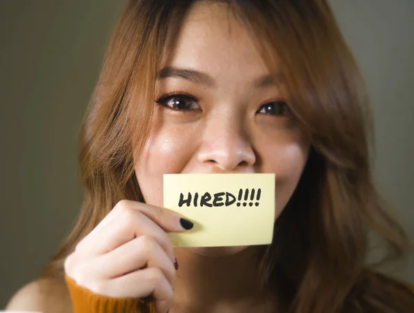 close up face portrait of young happy and beautiful Asian Korean student girl holding text message note with word hired handwritten excited about new job in successful work search concept