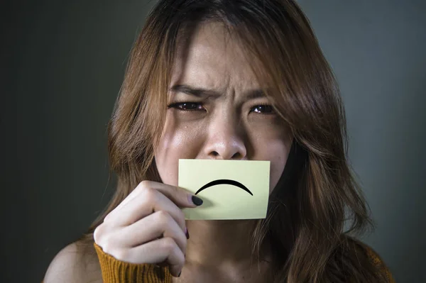 dramatic portrait of young overwhelmed and depressed Asian Korean student girl holding paper with sad mouth feeling desperate and frustrated suffering depression problem isolated on dark bg