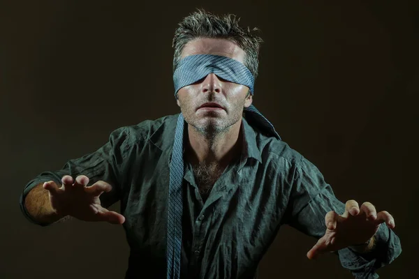 young lost and confused man blindfolded with necktie playing internet trend dangerous viral challenge with eyes blind acting doubtful guided by intuition isolated on dark background