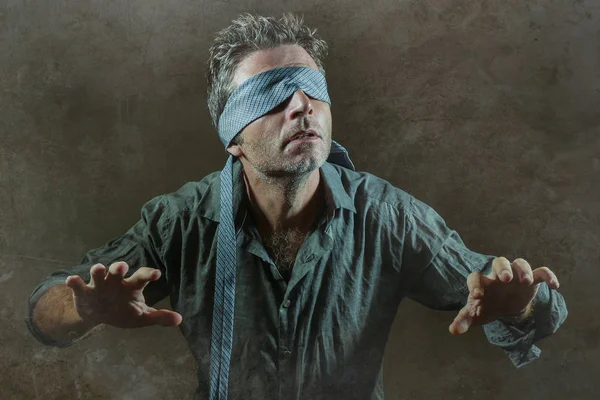 young lost and confused man blindfolded with necktie playing internet trend dangerous viral challenge with eyes blind acting doubtful guided by intuition isolated on dark background
