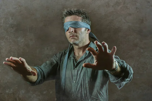 young lost and confused man blindfolded with necktie playing internet trend dangerous viral challenge with eyes blind acting doubtful guided by intuition isolated on dark background