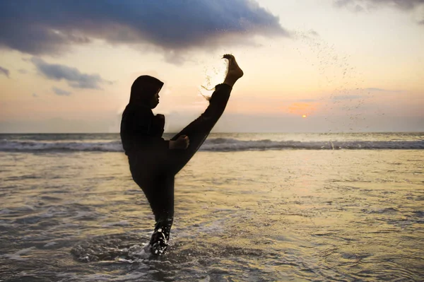 silhouette of young fit and aggressive Muslim woman covered in Islam hijab head scarf training martial arts karate kick attack and fitness workout at beautiful beach sunset in defense concept