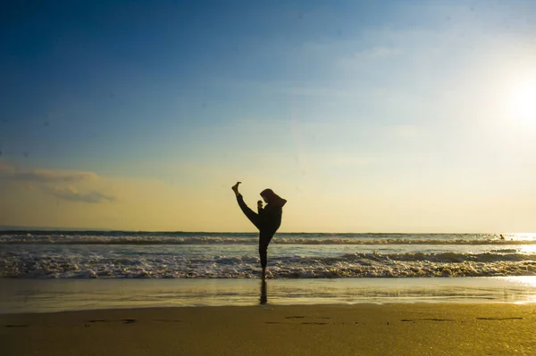 silhouette of young fit and aggressive Muslim woman covered in Islam hijab head scarf training martial arts karate kick attack and fitness workout at beautiful beach sunset in defense concept