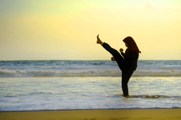 silhouette of young fit and aggressive Muslim woman covered in Islam hijab head scarf training martial arts karate kick attack and fitness workout at beautiful beach sunset in defense concept