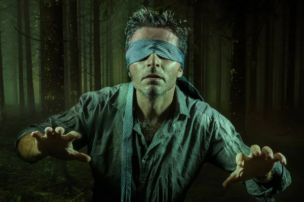 young confused and scared man blindfolded with necktie playing internet trend dangerous viral challenge with eyes blind lost in dark forest background guided by intuition