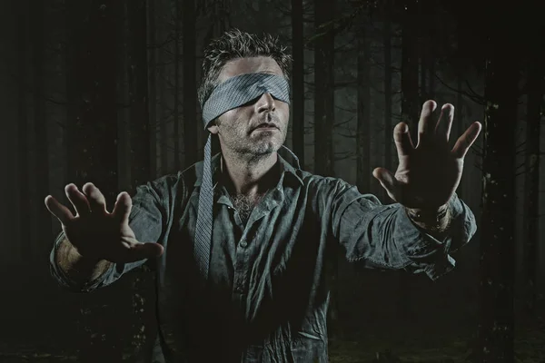 young confused and scared man blindfolded with necktie playing internet trend dangerous viral challenge with eyes blind lost in dark forest background guided by intuition