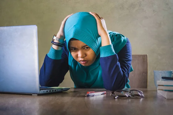 young stressed and overwhelmed Muslim student woman in Islam hijab head scarf  studying tired feeling overworked working with laptop computer and University books preparing exam