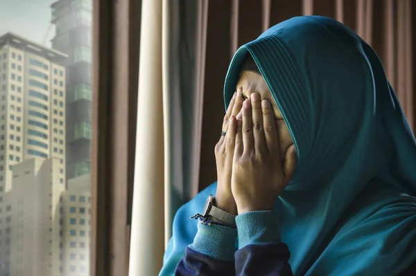 lifestyle portrait of young sad and depressed Muslim woman in Islam traditional Hijab head scarf at home window feeling unwell suffering depression crisis and anxiety problem crying helpless