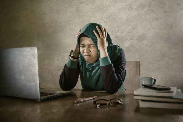 young stressed and overwhelmed Muslim student woman in Islam hijab head scarf  studying tired feeling overworked working with laptop computer and University books preparing exam