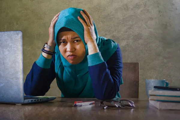 young stressed and overwhelmed Muslim student woman in Islam hijab head scarf  studying tired feeling overworked working with laptop computer and University books preparing exam