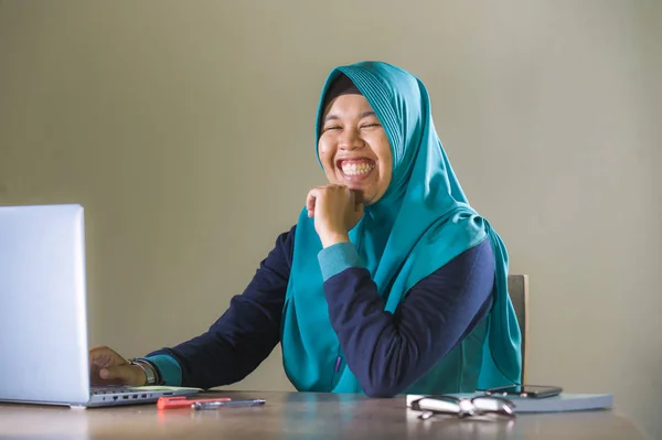 young happy and successful Muslim student woman in traditional Islam hijab head scarf working on desk studying with laptop computer and textbook smiling confident and cheerful