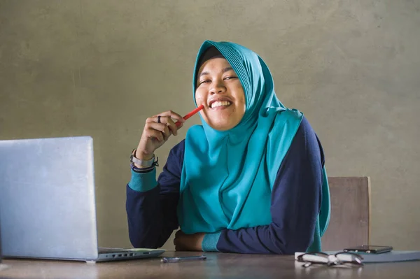 young happy and successful Muslim student woman in traditional Islam hijab head scarf working on desk studying with laptop computer and textbook smiling confident and cheerful
