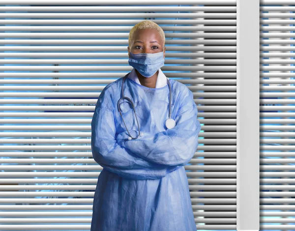 young attractive and confident black African American medicine doctor wearing face mask and blue scrubs standing corporate isolated at hospital room with Venetian blinds background