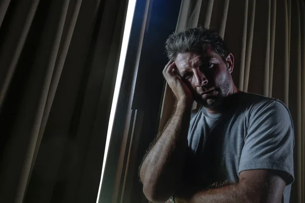 dramatic light indoors portrait of young sad and depressed attractive man at home looking through room window thoughtful and pensive lost in pain broken heart suffering depression problem