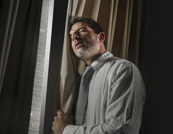 late night office portrait of young stressed and overwhelmed businessman working under pressure feeling depressed and worried suffering anxiety crisis leaning on window in corporate job problem