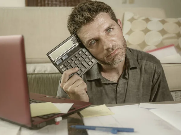 young stressed and worried man at home living room using calculator and laptop doing domestic accounting paperwork feeling overwhelmed on money tax expenses and financial status