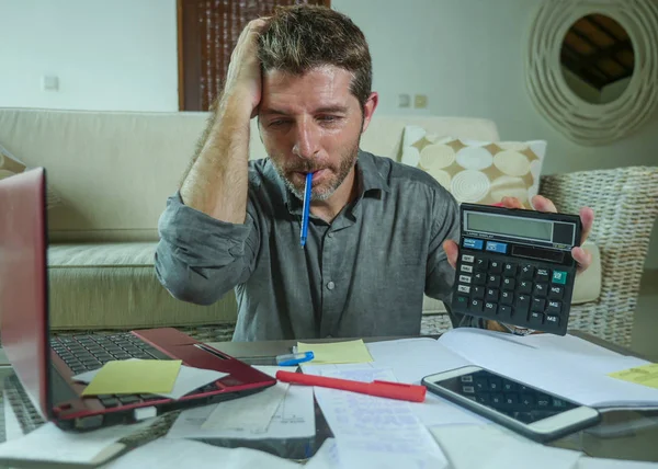 young stressed and worried man at home living room using calculator and laptop doing domestic accounting paperwork feeling overwhelmed on money tax expenses and financial status