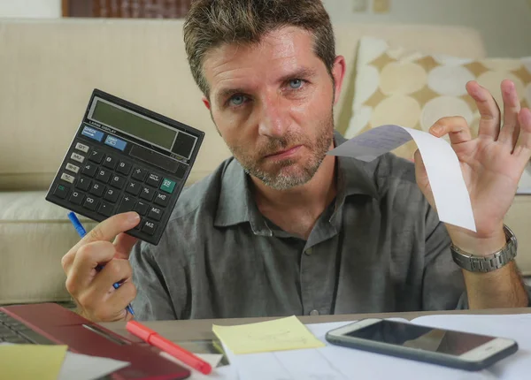 young stressed and worried man at home living room using calculator and laptop doing domestic accounting paperwork feeling overwhelmed on money tax expenses and financial status
