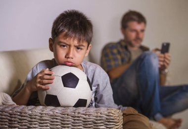 young sad and bored child at home couch feeling frustrated and unattended waiting his father for playing football while man networking on mobile phone as internet addict father neglecting son